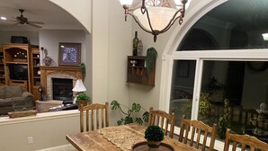 A dining area with a wooden table, chairs, and a chandelier. There is a fireplace with a clock and a television in the background.
