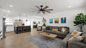 A spacious living room with a sofa, coffee table, and a ceiling fan. There is a kitchen area with a dining table and chairs.