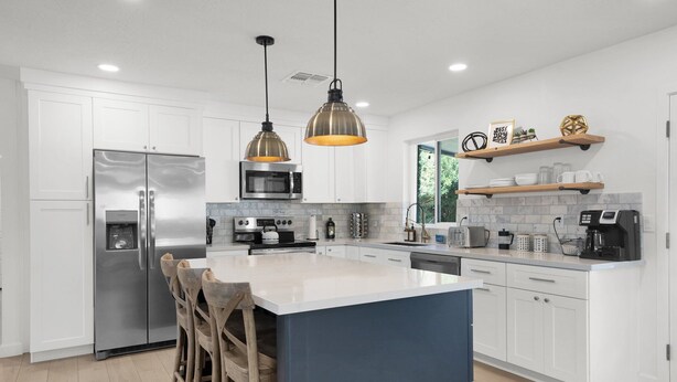 A modern kitchen with white cabinets, stainless steel appliances, a large island, hanging pendant lights, and wooden chairs.