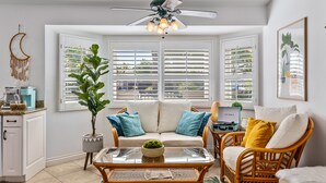Un salon chaleureux avec un canapé, une table basse et des fauteuils en osier, un ventilateur de plafond et une vue dégagée sur l’extérieur grâce à de grandes fenêtres.