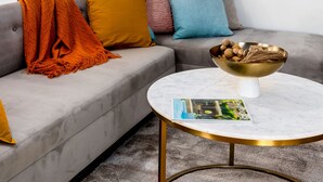 Un salon moderne avec un canapé gris, des coussins colorés, une table basse ronde sur laquelle repose un magazine, et trois tableaux encadrés accrochés au mur.