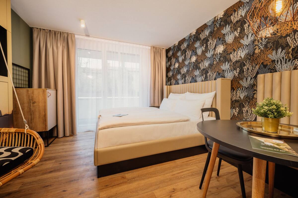 Habitación de hotel con cama, mesa de comedor, una silla colgante y una pared con diseño de hojas.