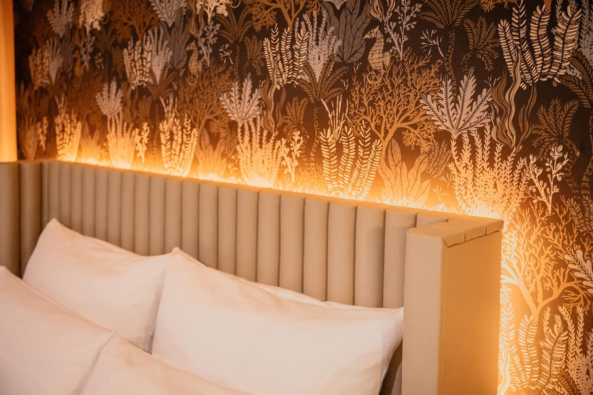 Una cama con cabecera y almohadas, frente a una pared con papel tapiz de hojas.