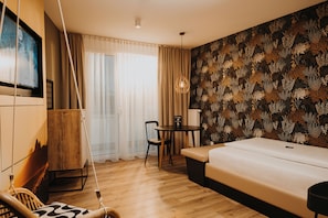 A modern hotel room with a wooden floor, a white sofa, a dining table with chairs, a TV mounted on the wall, and a patterned wallpaper.