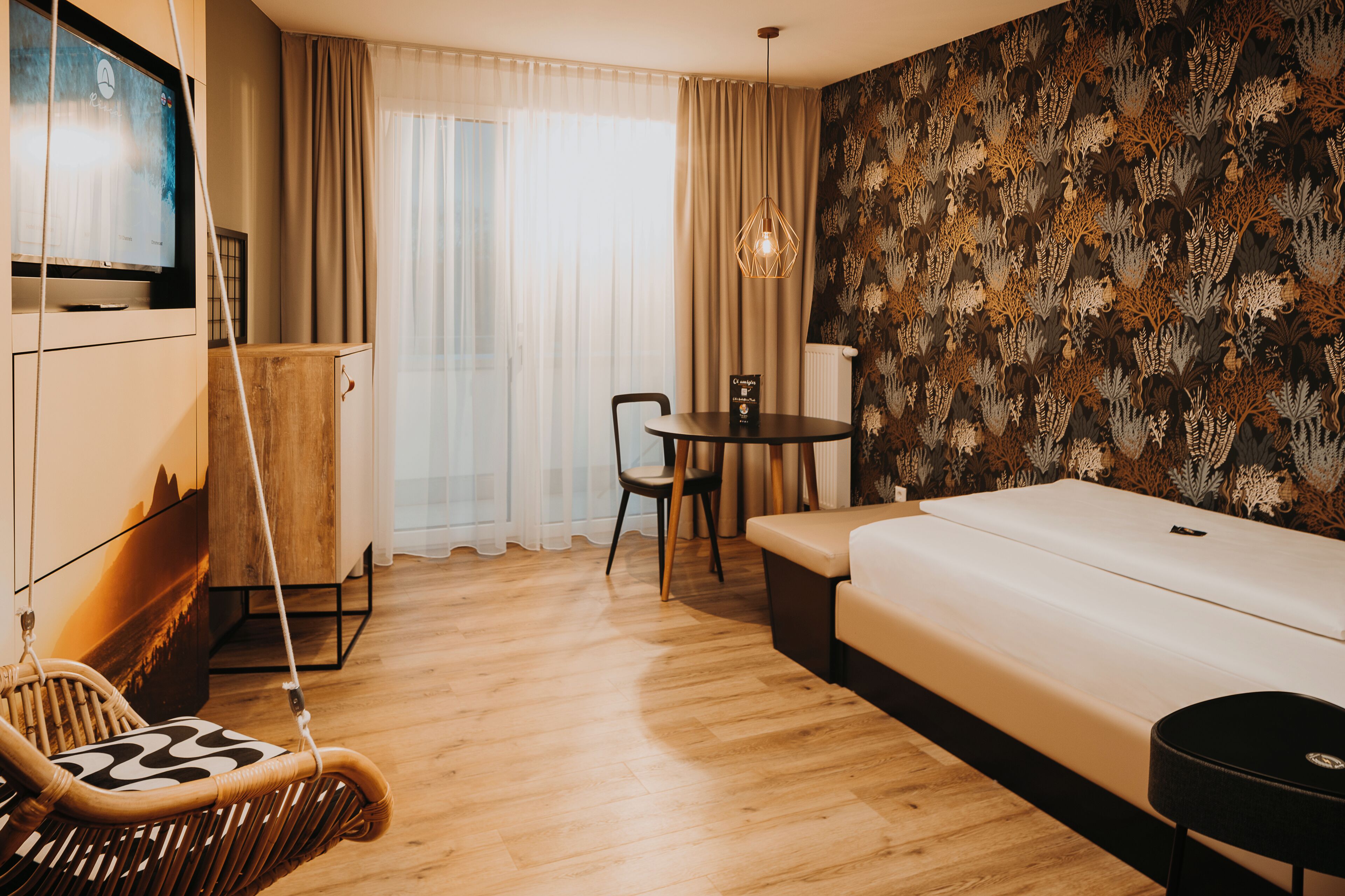 Ein modernes Hotelzimmer mit Holzboden, einer Couch, einem Esstisch mit Stühlen, einem Hängesessel und einem gemusterten Wandteppich.