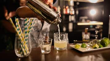 En bartender hælder drik i et glas, med en bakke limefrugter og en flaske i baggrunden.