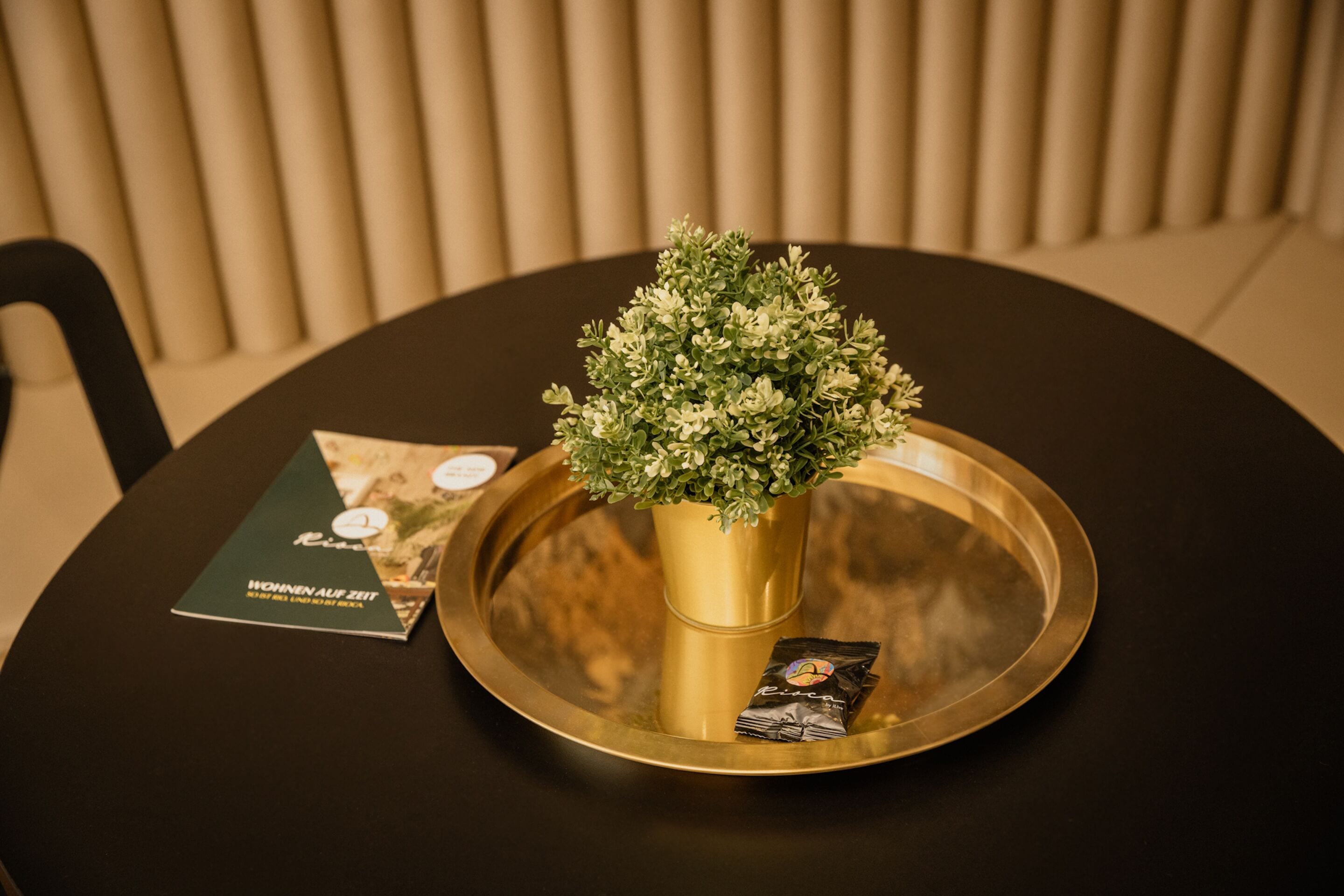 Une table ronde sur laquelle se trouvent un plateau doré, une petite plante en pot et une brochure.