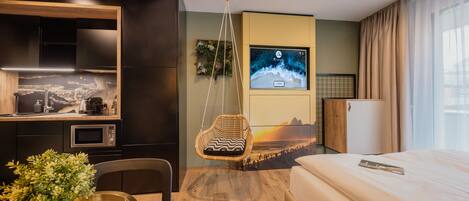 A modern hotel room with a bed, a hanging chair, a TV, a kitchen area, and a view of the cityscape.