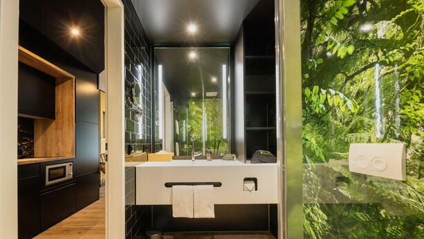 A modern bathroom with a large mirror, a sink, and a shower. There is a wooden cabinet and a trash can. - Rioca Stuttgart Posto 4 (Stuttgart)