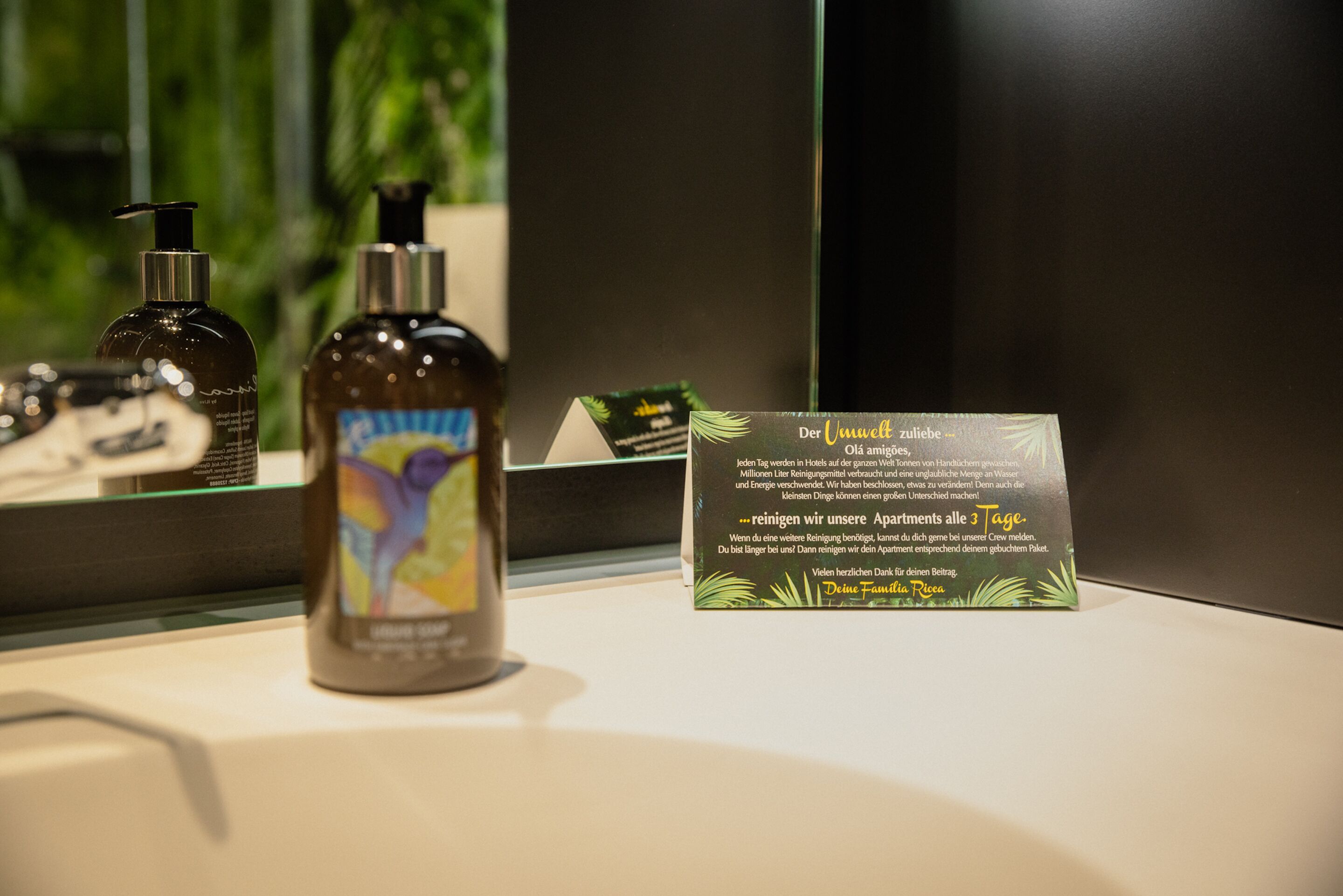 Una encimera de baño con una botella de jabón líquido y un cartel con texto en alemán.