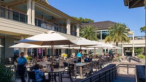 An outdoor patio area with tables and chairs, umbrellas, and a building with a balcony in the background. - Spectacular Studio in the Heart of Harbour Town overlooking Harbour & Lighthouse. Dine, Shop, Relax. (Hilton Head Island)