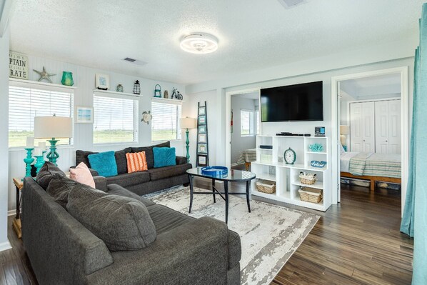 A living room with a sofa, a coffee table, and a TV. There is a bookshelf and a window with blinds.