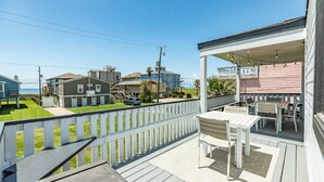Une terrasse en bois, dotée de mobilier d’extérieur, d’une rambarde blanche et offrant une vue sur les maisons voisines et une plage.