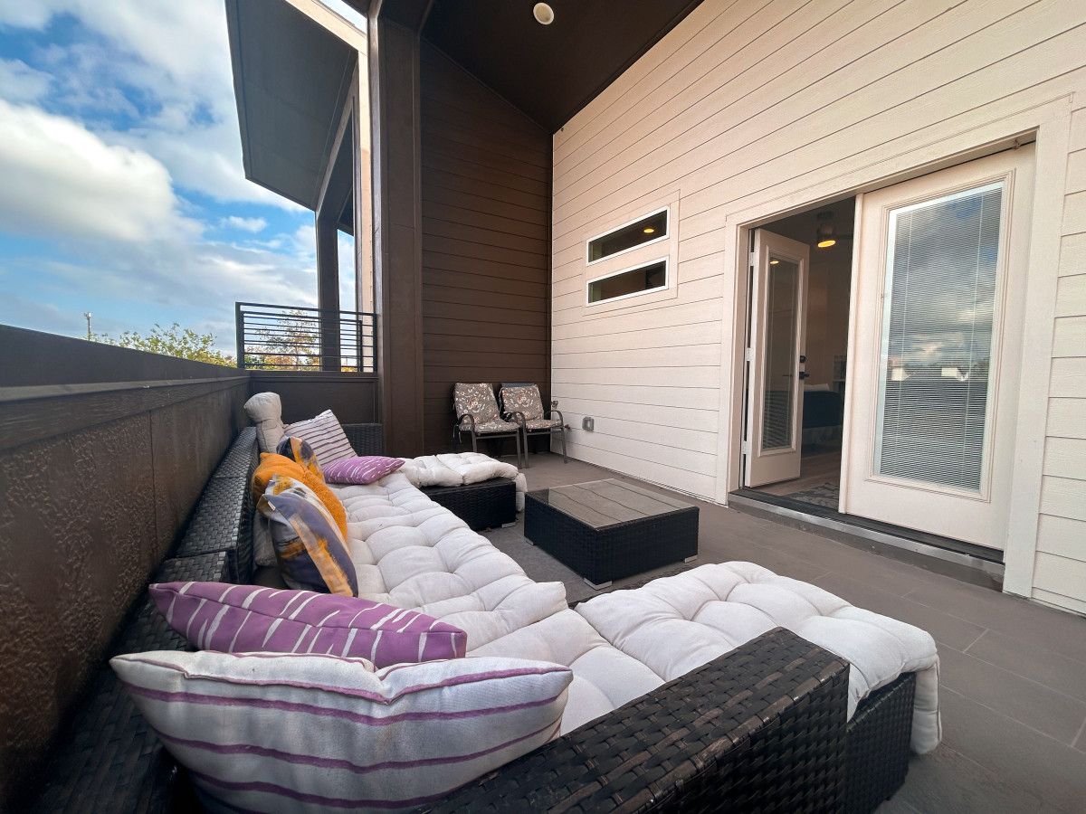 A covered outdoor seating area with cushioned benches and chairs, a sliding glass door, and a view of the sky.