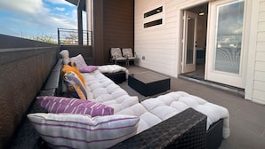 A covered outdoor seating area with cushioned benches and chairs, a sliding glass door, and a view of the sky. - 4 BR Townhome w/ Rooftop + Skyline View (Houston)
