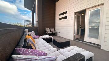 A covered outdoor seating area with cushioned benches and chairs, a sliding glass door, and a view of the sky.