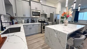 A modern kitchen with grey cabinets, a central island, stainless steel appliances, and a dining area with bar stools. - 4 BR Townhome w/ Rooftop + Skyline View (Houston)