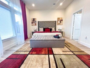 A bedroom with a large bed, a headboard, a bedside table, a nightstand, a window with curtains, and a door. - 4 BR Townhome w/ Rooftop + Skyline View (Houston)