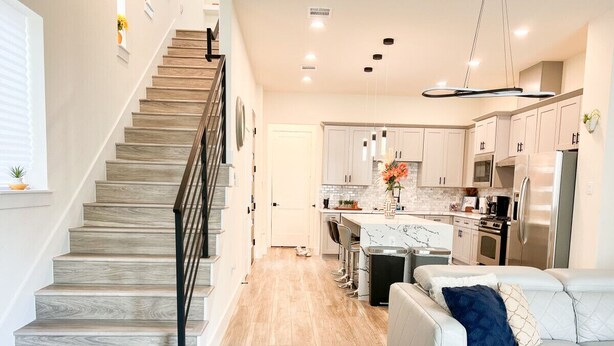 A modern kitchen with a central island, stainless steel appliances, and a staircase with a railing. - 4 BR Townhome w/ Rooftop + Skyline View (Houston)