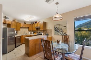 A modern kitchen with wooden cabinets, a stainless steel refrigerator, a glass dining table, and a view of mountains through the window.