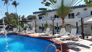 A swimming pool with lounge chairs and umbrellas, surrounded by palm trees and a building with blue accents. - Hotel Paraíso Kora (Aticama)