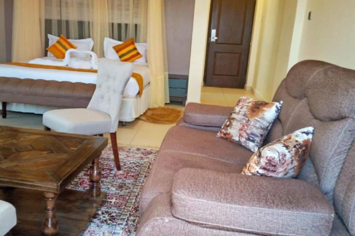 A spacious living room with a large sofa, a matching armchair, and a wooden coffee table. There is a bed with pillows and a nightstand in the background.
