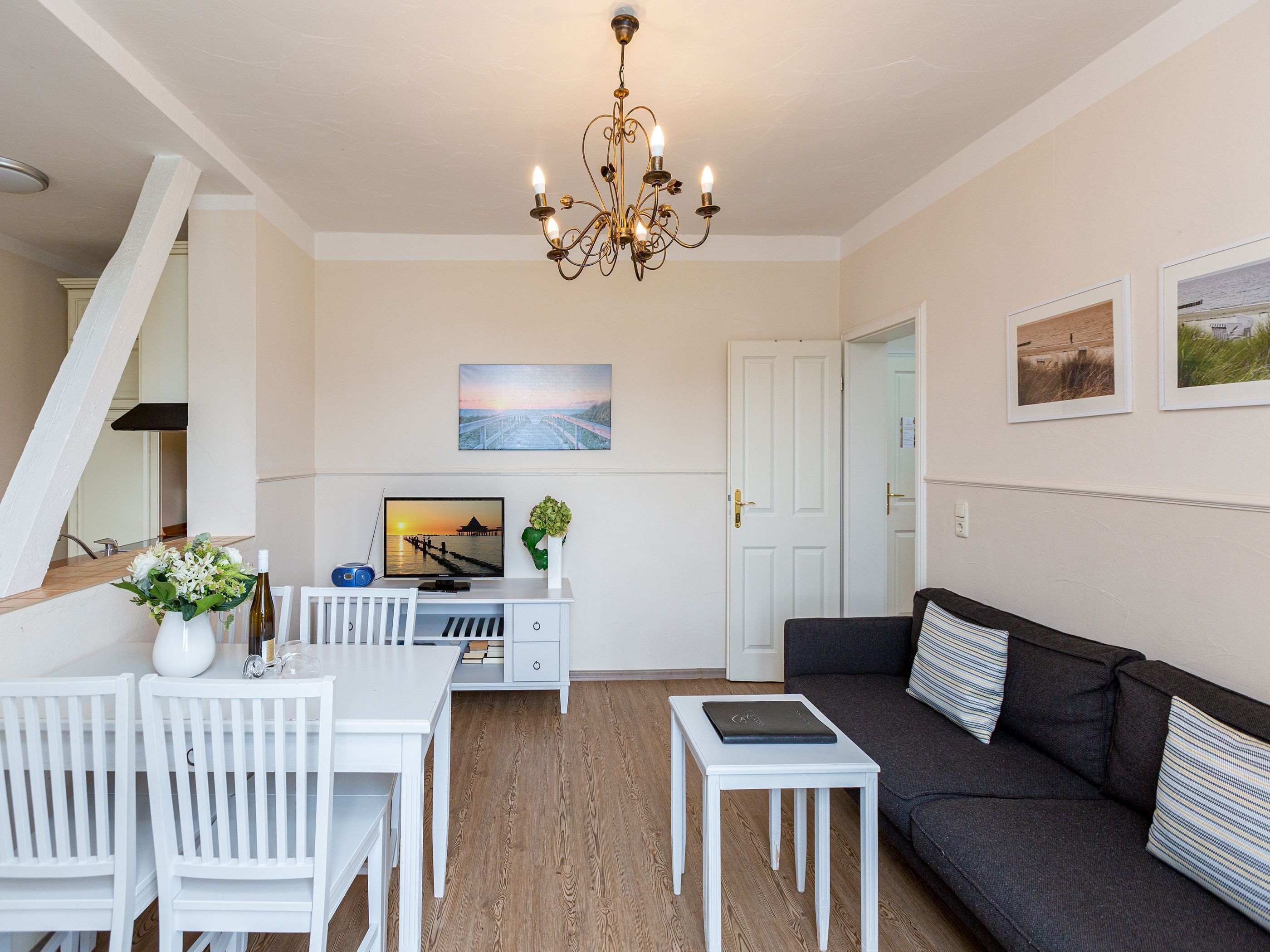Ein modernes Wohnzimmer mit einem weißen Esstisch, weißen Stühlen, einem dunkelgrauen Sofa und einem kleinen weißen Beistelltisch. Eine Kandelaber hängt von der Decke, ein Gemälde ist an der Wand und eine Tür führt in einen weiteren Raum.