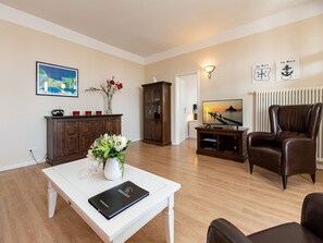 Un salon avec une table basse blanche, un fauteuil en cuir marron, un téléviseur, un meuble contenant des tasses rouges et un tableau encadré accroché au mur.