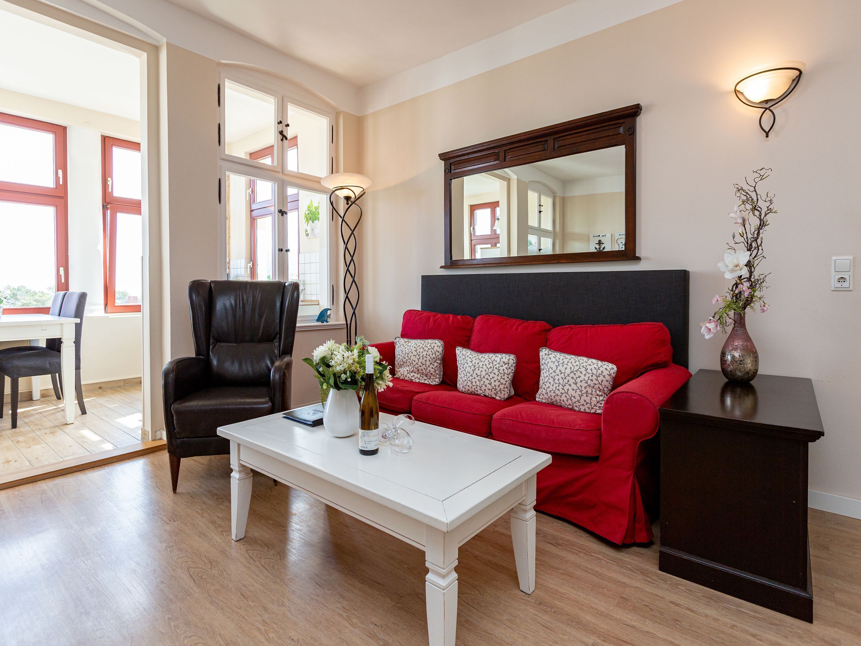 Una sala de estar con un sofá rojo, una mesa de centro blanca, un sillón de cuero negro, un armario de madera oscura y un espejo grande en la pared.
