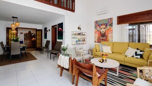Un amplio espacio habitable con un sofá amarillo, una mesa de comedor y sillas, una escalera a un nivel superior, y un cuadro colorido en la pared.