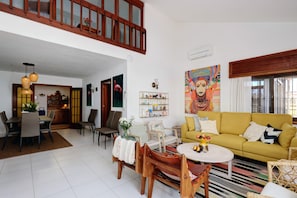 A spacious living area with a yellow sofa, dining table, and chairs, a staircase to an upper level, and a colorful painting on the wall.