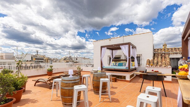 Terraza en la azotea con plantas en macetas, barriles de madera, taburetes blancos y una pequeña cocina al aire libre.