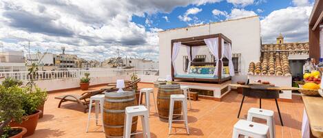 Terraza en la azotea con plantas en macetas, barriles de madera, taburetes blancos y una pequeña cocina al aire libre.