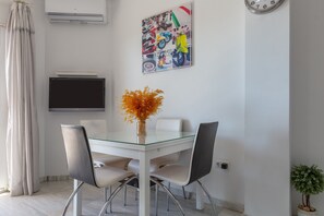 A dining area with a glass table, four chairs, a vase with orange flowers, a wall-mounted TV, and a poster with various images.