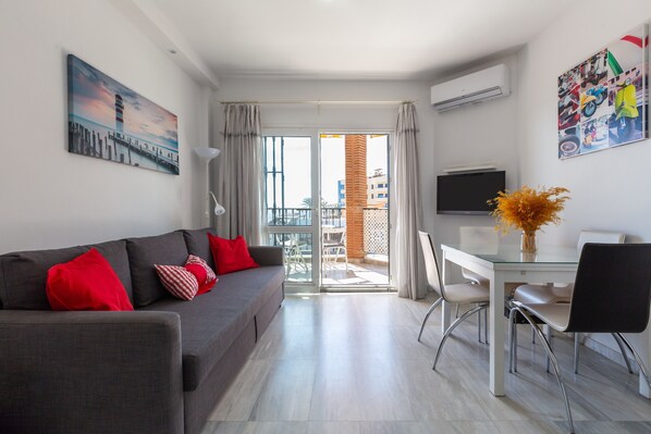A modern living room with a sofa, a dining table, chairs, a TV, and a balcony with a view.