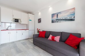 A modern kitchen with white cabinets, a microwave, and a red countertop. There is a gray sofa with red cushions and a painting of a pier on the wall.