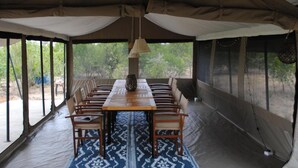A tented dining area with a long table and chairs, surrounded by windows offering a view of the outdoors.