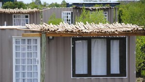 A small, single-story building with a simple, functional design. It features a large window with a black frame and a door with a grid pattern. The roof is covered with wooden beams.