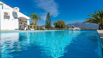 A clear blue pool with lounge chairs, a white building with a satellite dish, and a mountain view.