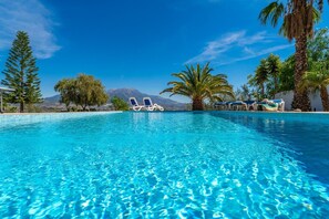 Piscina cristallina azzurra con lettini, palme e una montagna sullo sfondo.