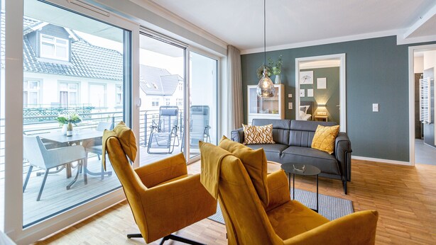 Ein modernes Wohnzimmer mit einer Couch, zwei Sesseln, einem Esstisch und Blick durch große Fenster.