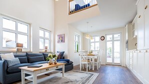 Ein großzügiger Wohnraum mit Sofa, Couchtisch und Essbereich mit Stühlen. Ein großes Fenster lässt viel Tageslicht herein und bietet einen Ausblick.