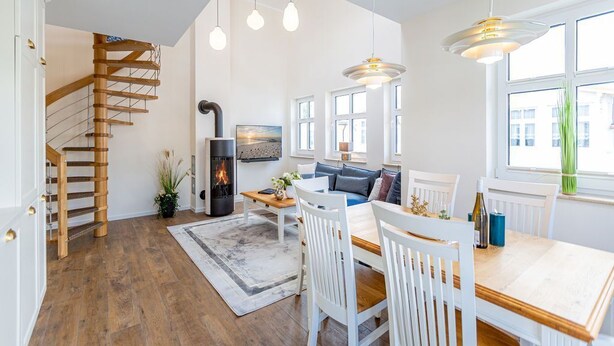 Ein modernes Wohnzimmer mit einer Holztreppe, einem Kamin, einem Essbereich mit weißen Stühlen und großen Fenstern.
