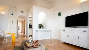 Un salon avec un canapé blanc, une table basse sur laquelle on peut voir un jeu d’échecs, un téléviseur, et une cuisine en arrière-plan.