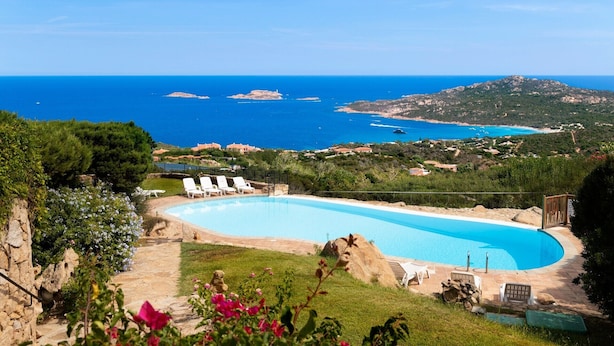 Une piscine bordée de chaises longues, un mur en pierre et une vue sur la mer et les collines.