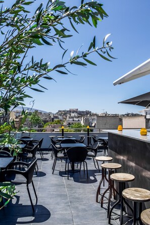 Uma área externa com mesas e cadeiras, vista para o horizonte urbano e um edifício com design moderno.
