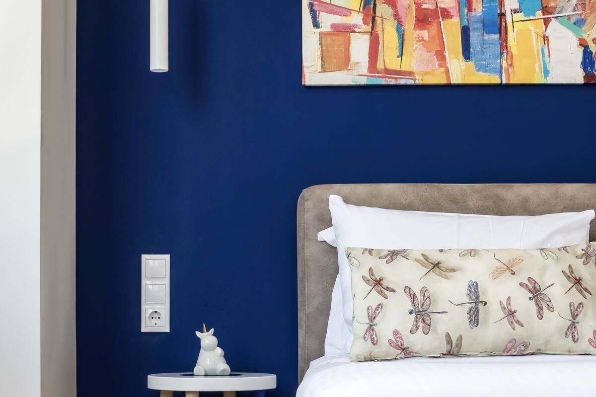 Un dormitorio con pared azul, una cama blanca con un cojín de libélula, una mesita blanca con una figurita de unicornio y un cuadro sobre la cama.