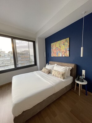 Un dormitorio moderno con una cama grande, vistas de la ciudad a través de una ventana, una pared de acento azul y un teléfono en la mesita de noche.