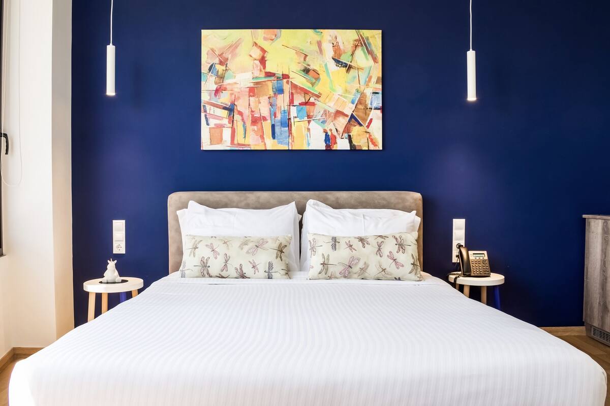 Una habitación de hotel con una cama grande, dos almohadas con un diseño de libélula, un cuadro abstracto colorido en la pared y un teléfono sobre una mesita de noche.