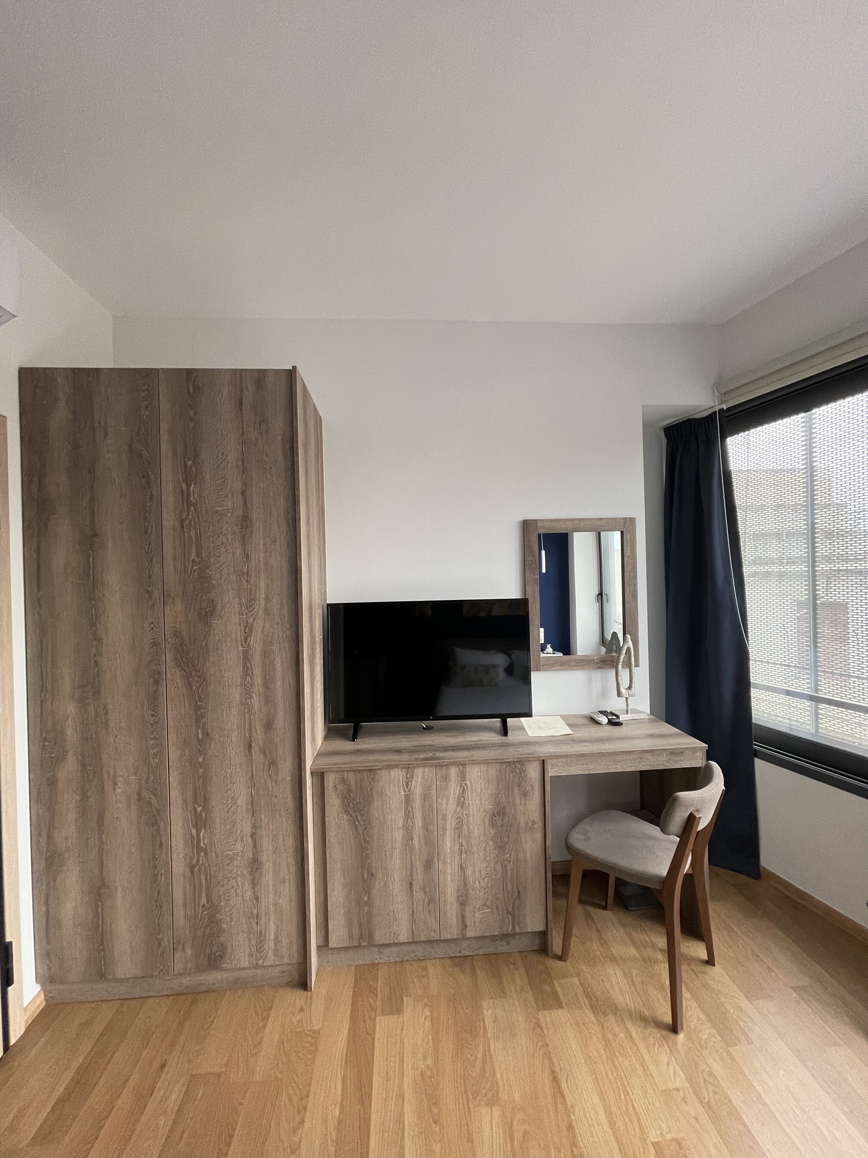 Ein modernes Zimmer mit Schreibtisch aus Holz, Fernseher, Sessel und einem großen Fenster mit Vorhängen.